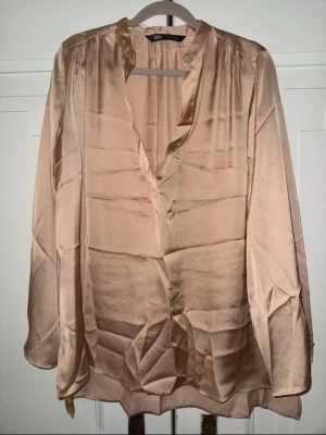 Champagnefärgad satinblus från ZARA - Snygg champagnefärgad blus i glansigt satinmaterial från ZARA. Blusen har v-ringning, avslappnad passform, kortare ärmar och något längre baktill. Blusen är något stor i storleken. Perfekt att styla med jeans eller kjol för en trendig look.