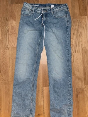 Blå raka jeans med snörning - Ett par ljusblå jeans med rak passform och låg midja. Jeansen har en enkel design med fem fickor och snörning i midjan istället för knapp. Tillverkade i klassiskt denimtyg som ger en avslappnad vibe.