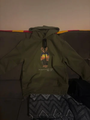 Olivgrön hoodie Polo bear Ralph Lauren - Snygg olivgrön hoodie från Polo Ralph Lauren med ikoniska Polo Bear-trycket på bröstet. Hoodien har huva och ribbade muddar. Materialet känns mjukt och skönt, perfekt för en avslappnad streetwear-look. Märkestagg kvar i halsen. NYPRIS: 2795kr. Använd en endaste gång, anledningen till försäljning är att den är liten. Det är storlek S men ifall du är över 175cm är den väldigt kort i midjan. Överlag en sjukt härlig hoodie