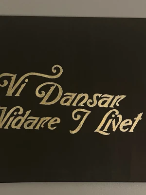 ”vi dansar vidare i livet” hov1 tavla 50x70 - Snygg svart tavla med guldfärgad text i snirkligt typsnitt där det står 'Vi Dansar Vidare I Livet'. Perfekt som väggdekoration för dig som gillar inspirerande citat och stilren inredning.