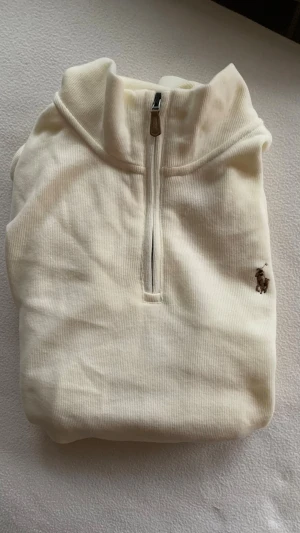 Beige halfzip från Ralph Lauren - Klassisk beige halvzip-tröja från Ralph Lauren med broderad logga på bröstet. Tillverkad i mjukt ribbat material och har hög krage med dragkedja. Perfekt för en clean och stilren look.
