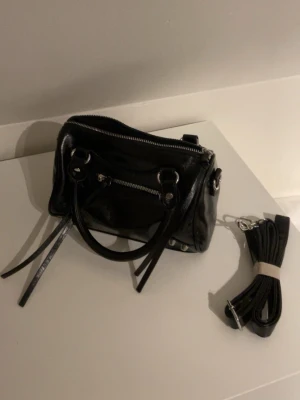 Svart handväska i skinn med dragkedja - Snygg svart handväska i skinn med silverfärgade detaljer och dragkedja upptill. Väskan har både handtag och en avtagbar axelrem som kan fästas med karbinhakar. Perfekt för dig som vill ha en stilren och praktisk väska till vardagen.