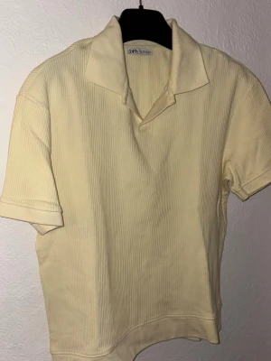 Gul  - Säljer en ljusgul ribbad polotopp från Zara. Toppen har korta ärmar, klassisk krage och är tillverkad i ett mjukt material som känns skönt mot huden. Perfekt att styla med jeans eller kjol för en clean look.