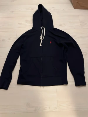 Marinblå hoodie från Polo Ralph Lauren - Snygg marinblå hoodie med dragkedja från Polo Ralph Lauren nypris 1600kr. Klassisk design med vit dragsko i huvan och röd logga broderad på bröstet. Mjuk bomullskänsla och två fickor framtill. Perfekt för en avslappnad och stilren look.
