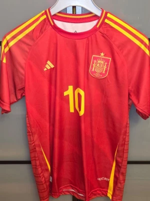 Spaniens matchtröja Lamine Yamal #10 - Röd matchtröja från Adidas för Spaniens landslag med gula detaljer och nummer 10. På bröstet finns landslagets emblem och Adidas-logga, på ryggen står Lamine Yamal och siffran 10. Tillverkad i funktionsmaterial HEAT.RDY för bästa komfort under match. Den är helt ny aldrig använd men den är bara tvättad. Priset är inte hugget i sten