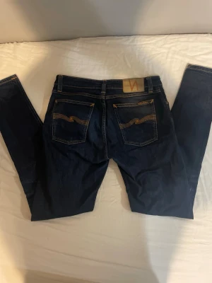 Mörkblå jeans från Nudie Jeans - Säljer ett par mörkblå jeans från Nudie Jeans med klassiska kontrastsömmar och ikonisk broderad bakficka. Jeansen har fem fickor och en slim passform som sitter snyggt. Perfekta för dig som gillar stilrena och tidlösa jeans.