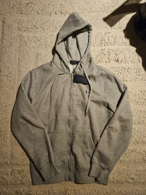 Grå zip-up Ralph Lauren - Grå zip-up från Ralph Lauren i storlek L men passar M. Lappar kvar. Dragskon har en defekt.