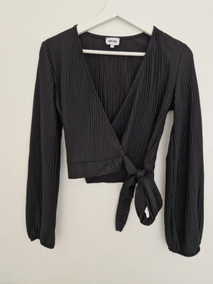 Svart omlottopp från Bubbleroom XS - Säljer en svart omlottopp från Bubbleroom i storlek XS. Toppen har långa ärmar, plisserat tyg och knyt i sidan. Tillverkad i 100% polyester och har en croppad passform med v-ringad hals. Perfekt att styla med höga jeans eller kjol.