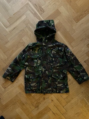 Camouflage jacka - Säljer en grön och brun camouflage jacka med huva och flera praktiska fickor framtill. Jackan har dragkedja och knappar, samt justerbar midja.