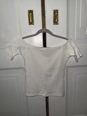 Vit ribbad offshoulder topp NA-KD - Trendig vit ribbad topp från NA-KD med offshoulder-design och korta ärmar. Tillverkad i mjuk bomullsmix som sitter snyggt och bekvämt. Perfekt att styla med jeans eller kjol för en clean look.