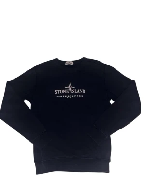  Stone Island tröja - Mörkblå sweatshirt från Stone Island med vit logga och texten 'Otherside Voyager 7116' på bröstet. Klassisk rund halsringning och ribbade muddar vid ärmslut och nederkant. Perfekt för en clean och stilren look.