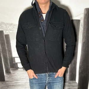 Svart overshirt med knappar - Stilren svart overshirt med knäppning framtill och två bröstfickor med lock. Klassisk krage och långärmad modell som passar perfekt över en hoodie eller skjorta. Enkel och mångsidig design som funkar till det mesta.