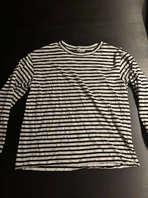 Randig långärmad topp från Lindex - En klassisk svartvit randig långärmad topp från Lindex. Toppen har rund halsringning och är gjord i mjuk bomull, perfekt för en chill och stilren look. Passar till allt och är superenkel att matcha med jeans eller kjol.