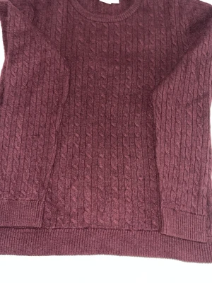 Vinröd kabelstickad tröja - Säljer en vinröd kabelstickad tröja med rund halsringning och ribbade muddar vid ärmslut och nederkant. Tröjan har ett klassiskt flätmönster och är perfekt för dig som gillar en stilren och mysig look.