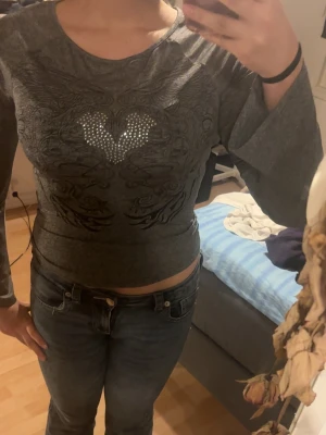 Mörkgrå croppad topp med strasshjärta - Säljer en mörkgrå croppad topp med lång ärm och ett coolt tribal-inspirerat mönster framtill. Toppen har ett hjärta i strass mitt på bröstet som ger extra bling. Osäker vart den är ifrån men tror de är antigen Shein eller temu , aldrig använd .