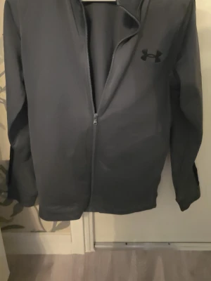 Svart träningsset från Under Armour - Säljer ett svart träningsset från Under Armour med ziptröja och matchande byxor. Setet har diskret logga på både jacka och byxa, samt svarta sidopaneler på byxorna. Perfekt för gymmet eller löprundan, tillverkat i ett stretchigt funktionsmaterial.