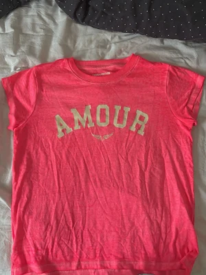 Rosa zadig volitaire t-shirt - Cool rosa T-shirt med texten 'AMOUR' i vitt framtill och en liten vingdetalj under från zadig volitaire. T-shirten har rund halsringning och korta ärmar. Perfekt för en avslappnad och trendig look.