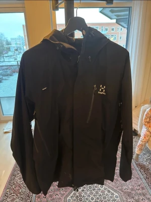 jacka från Haglöfs - Svart jacka från Haglöfs med huva och dragkedjor framtill. Jackan har en stilren design med logga på bröstet och praktiska fickor med dragkedja. Perfekt för regniga dagar och outdoor-äventyr. Liten fläck nertill som man ser på bild annats toppen 