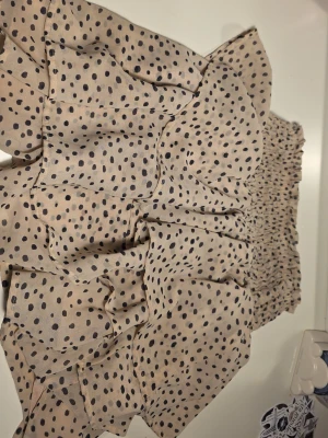 Beige prickig volangkjol - Supersöt kort beige kjol med svarta prickar och flera volanger. Kjolen har smock i midjan för en bekväm passform och är gjord i ett lätt och luftigt material. Perfekt för dig som gillar en lekfull och trendig stil.