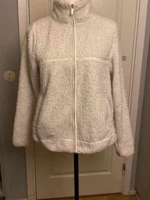 Vit teddyjacka med dragkedja - Mysig vit teddyjacka med hög krage och hel dragkedja framtill. Jackan har lång ärm och två sidofickor. Perfekt för kalla dagar när du vill vara både varm och stilren. Materialet är fluffigt och ger en avslappnad look.
