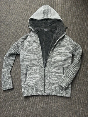 Grå stickad hoodie med dragkedja - Sjukt fet gråmelerad stickad hoodie med dragkedja och två fickor med blixtlås. Insidan är fodrad med mjuk svart fleece för extra värme. Tröjan har ribbade muddar och en stor huva, perfekt för kyliga dagar. Jätte fint skick, tveka inte på att höra av er vid frågor eller funderingar! 