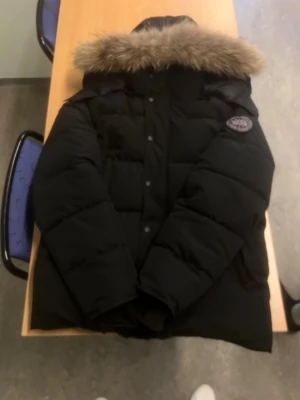 Svart dunjacka från Canada Goose - Säljer en svart dunjacka från Canada Goose med stor, fluffig fuskpäls runt huvan och klassisk patch på ärmen. Jackan har quiltad design, dragkedja och knappar framtill samt lång ärm. Perfekt för kalla vinterdagar.