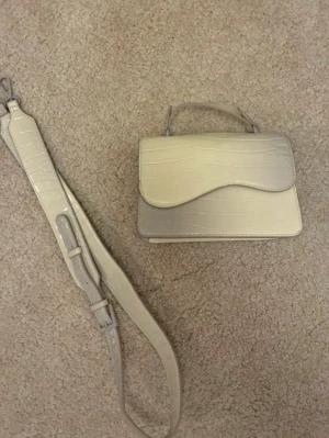 Beige croco handväska med axelrem - Snygg beige handväska i croco-mönstrat syntetmaterial. Väskan har ett topphandtag och en avtagbar, justerbar axelrem. Perfekt för dig som vill ha en stilren och trendig accessoar till din outfit.