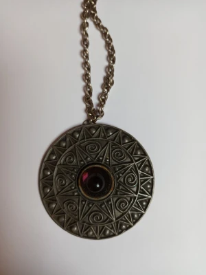  vintage halsband med ametist och mönster - Unikt halsband med en stor, rund medaljong i antik silverfärg. Medaljongen har ett detaljerat mönster och en lila sten troligtvis ametist sten i mitten. Kedjan är grov.