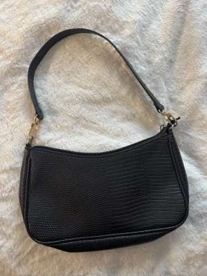 Svart enkel axelväska i skinn. - Svart enkel axelväska i skinn. Väskan har en enkel, avlång form och stängs med dragkedja. Små guld grejer vid sidorna.