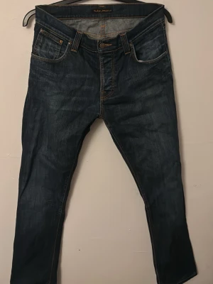 Mörkblå jeans från Nudie Jeans - Säljer ett par mörkblå jeans från Nudie Jeans med klassisk femficksdesign och kontrastsömmar. Jeansen har raka ben och snygga detaljer på bakfickorna. Inga defekter, står att dem är 31/32 men skulle mer säga 30/32