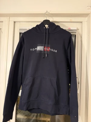 Marinblå hoodie från Tommy Hilfiger - Snygg marinblå hoodie från Tommy Hilfiger med klassisk logga och text på bröstet. Hoodien har huva med dragsko och en stor magficka framtill. Saknas en metallbit på slutet av snöret till luvan, se bild. Annars i bra skick. Tillverkad i mjukt bomullsmaterial och har ribbade muddar vid ärmslut och nederkant.