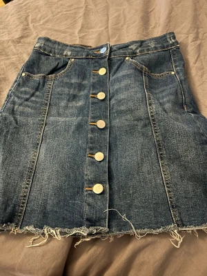 Jeanskjol med knappar framtill - Snygg jeanskjol i blå denim med fransig nederkant och dekorativa guldknappar längs hela framsidan. Knappt använd därför jag säljer den💙passar många då den är väldigt stretchy