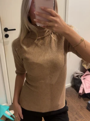 Beige ribbstickad polotröja - Snygg beige ribbstickad polotröja med halvlånga ärmar och hög krage. Tröjan har en figurnära passform och är perfekt att styla med jeans eller kjol. Mjuk och skön känsla i materialet.
