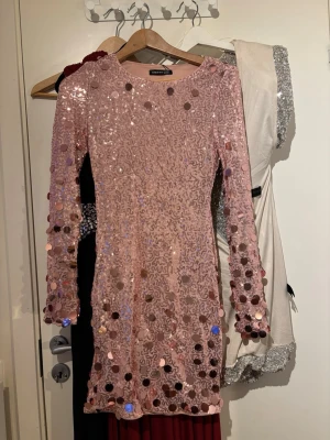 Rosa paljettklänning från Bubbleroom - Glittrig rosa kortklänning från Bubbleroom med långa ärmar och rund halsringning. Klänningen är täckt av paljetter och stora dekorativa paljettdetaljer som ger en festlig look. Perfekt för dig som vill sticka ut.