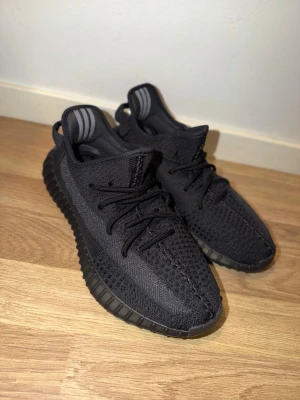 Oanvända Svarta adidas Yeezy Boost 350 V2 - Säljer ett par svarta adidas Yeezy Boost 350 V2 sneakers med stickad ovandel och klassisk ribbad sula. Skorna har snörning och diskreta ränder på hälen. Perfekta för dig som gillar streetwear och vill ha något stilrent och bekvämt.
