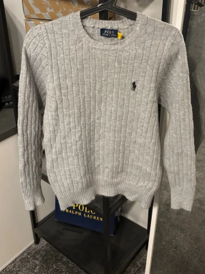 Grå kabelstickad tröja Polo Ralph Lauren - Kabelstickad grå tröja från Polo Ralph Lauren med rund halsringning och broderad logga på bröstet. Tröjan har långa ärmar och ribbade muddar. Perfekt för lager-på-lager och klassisk stil.