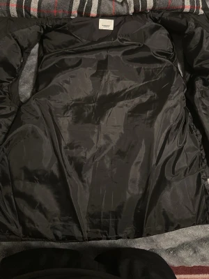 Svart jacka med rutigt foder - Stilren svart jacka med klassiskt rutigt foder i grått, rött och vitt. Jackan har en slät yta och är helfodrad, perfekt för dig som vill ha en tidlös look. Passar till många olika outfits och har en medellång passform.