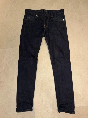 Mörkblå jeans från J.Lindeberg - Snygga mörkblå jeans från J.Lindeberg med klassisk femficksdesign och kontrastsömmar. Jeansen har en smal passform och är tillverkade i slitstarkt denimtyg. Perfekta för dig som gillar stilrena och tidlösa plagg.