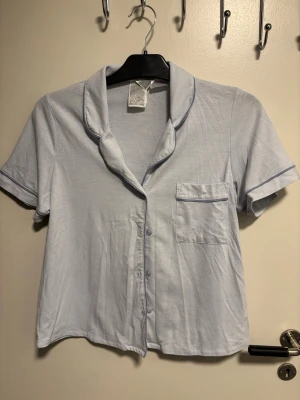 Ljusblå kortärmad pyjamasskjorta - Ljusblå kortärmad pyjamasskjorta med klassisk krage och knappstängning framtill. Skjortan har en bröstficka med diskret blå kant och är gjord i ett mjukt, tunt material som känns skönt mot huden. Perfekt för mysiga kvällar hemma.