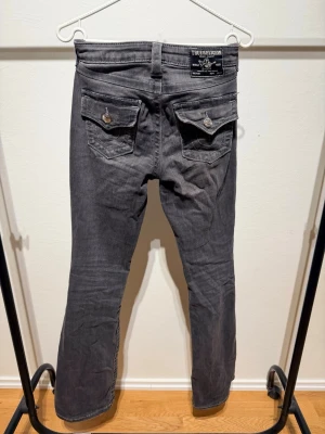 Grå jeans från True Religion - Snygga grå jeans från True Religion med utsvängda ben modell Becca,och klassiska kontrastsömmar. Jeansen har två fickor med lock och knapp bak samt femficksmodell fram. Tillverkade i mjukt denimtyg som ger en avslappnad vibe.