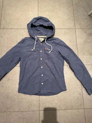 Sjukt snygg Hollister tröja  - Den är märkt som storlek L men skulle säga att den passar M bättre. Snygg blå overshirt från Hollister med huva och vita snören. Skjortan har två bröstfickor med knappar och klassisk knappstängning framtill. Perfekt för en avslappnad och trendig stil.
