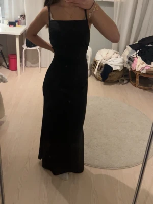 Svart långklänning med smala axelband - Stilren svart långklänning med smala axelband och rak halsringning. Klänningen har en figurnära passform och är perfekt för dig som gillar en enkel men elegant look. Materialet ser ut att vara mjukt och följsamt, vilket ger en snygg siluett.