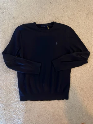 Mörkblå Stickad Tröja Polo Ralph Lauren Kashmir - Mörkblå stickad tröja från Polo Ralph Lauren med rund halsringning och diskret broderad logga på bröstet. Tröjan har långa ärmar och är tillverkad i mjuk bomull. Perfekt för en stilren och klassisk look.