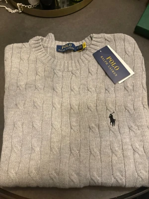 Grå kabelstickad tröja Polo Ralph Lauren - Snygg grå kabelstickad tröja från Polo Ralph Lauren med rund halsringning och klassisk broderad logga på bröstet. Tröjan har ribbade muddar och är tillverkad i mjukt material som passar perfekt till lager på lager. Osäker på äkthet då jag fick den som present