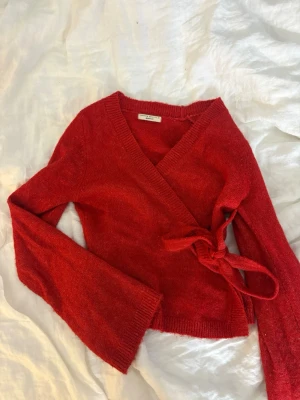 Röd omlottkofta från Gina Tricot - Snygg röd stickad omlottkofta från Gina Tricot med knytband i sidan och ribbade muddar. Koftan har en djup v-ringning och är tillverkad i ett mjukt, fluffigt material som känns lyxigt. Perfekt för dig som vill ha en trendig och bekväm look.