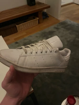 Vita adidas Stan Smith sneakers - Klassiska vita adidas Stan Smith sneakers med snörning och platt sula. Skorna har en stilren design med perforerade ränder på sidorna och adidas-logga på hälen. Tillverkade i skinn för en clean look.