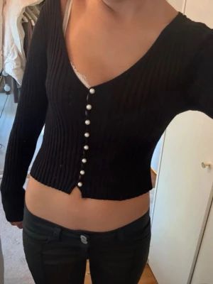 Svart ribbad kofta med pärlknappar - Trendig svart ribbstickad kofta med djup v-ringning och snygga pärlknappar framtill. Kort, figurnära modell med långa ärmar. 
