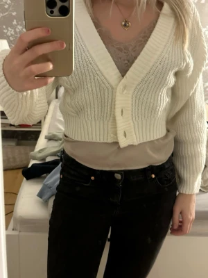 Vit cropped kofta med knappar - Säljer en vit, ribbstickad cropped kofta med v-ringning och stora knappar framtill. Koftan har långa ärmar och en bred ribbad kant nedtill. Perfekt att slänga över ett linne eller t-shirt för en trendig lager-på-lager-look.