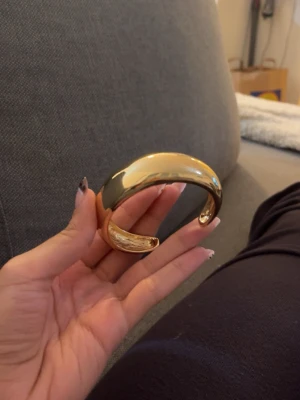 Bredt guldpläterat armband - Säljer ett chunky, guldpläterat armband med öppen design. Armbandet har en slät, glansig yta och en rundad form som ger en modern och stilren look. Perfekt statement-piece för dig som gillar att sticka ut med accessoarer.