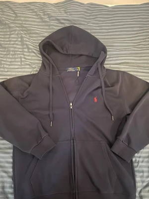 Mörkblå hoodie från Polo Ralph Lauren - Snygg mörkblå hoodie från Polo Ralph Lauren med dragkedja och huva. Klassisk röd logga broderad på bröstet och justerbara snören i huvan. Tillverkad i mjukt bomullsmaterial och har ribbade muddar vid ärmslut och nederkant. Perfekt för en avslappnad och stilren look.